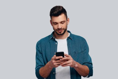 Beyaz genç adam akıllı telefonunu kullanarak ve gri arka plan karşı dururken gülümseyen