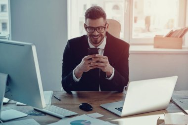 Yakışıklı genç adam akıllı telefonunu kullanarak ve ofiste otururken gülümseyen resmi kıyafeti