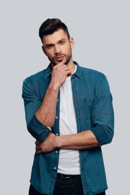 büyüleyici genç adam kameraya bakıyor ve gri arka plan aleyhine dururken çene üzerinde el tutmak