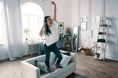 Deliriyorum. Evde kanepede dans ederken kulaklık takan ve gülümseyen çekici genç kadın tam uzunlukta