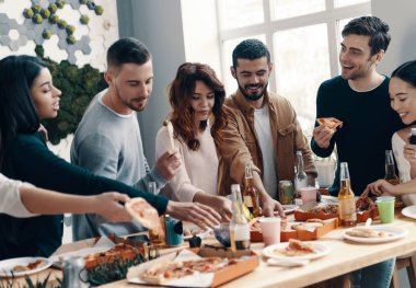 Sıcak pizzanın tadını çıkarıyorum. Gündelik giyimli bir grup genç, içeride akşam yemeği partisi verirken pizza yiyip gülümsüyor        
