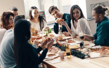 Gündelik giyimli bir grup genç, içeride akşam yemeği partisi verirken pizza yiyip gülümsüyor               