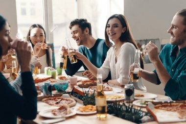 Harika vakit geçiriyor. Gündelik giyimli bir grup genç, içeride akşam yemeği partisi verirken pizza yiyip gülümsüyor