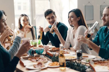 Rahatlatıcı bir akşam. Gündelik giyimli bir grup genç, içeride akşam yemeği partisi verirken pizza yiyip gülümsüyor