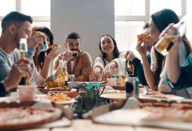 Arkadaşlarla daha çok gül. Gündelik giyimli bir grup genç, içeride akşam yemeği partisi verirken pizza yiyip gülümsüyor