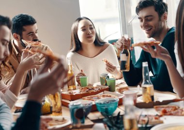 Harika vakit geçiriyor. Gündelik giyimli bir grup genç, içeride akşam yemeği partisi verirken pizza yiyip gülümsüyor 