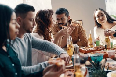 Lezzetli yemekleri paylaşmak. Gündelik giyimli bir grup genç, içeride akşam yemeği partisi verirken pizza yiyip gülümsüyor
