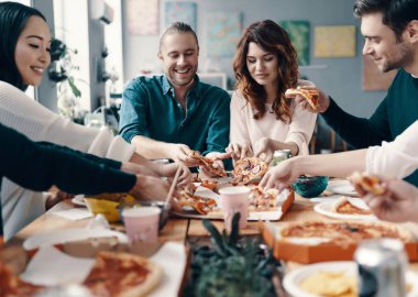 Pizza severler. Gündelik giyimli bir grup genç, içeride akşam yemeği partisi verirken pizza toplayıp gülümseyerek