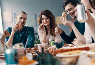 Tüm endişeleri geride bırak. Gündelik giyimli bir grup genç, içeride akşam yemeği partisi verirken pizza yiyip gülümsüyor           