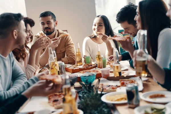 友達との生活は良い カジュアルウェアの若者のグループは 屋内でディナーパーティーを持っている間 ピザを食べて笑顔を身に着けている — ストック写真