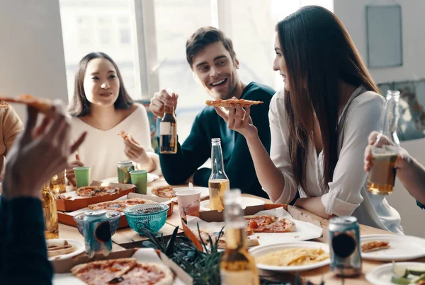 良い食べ物と良い友人 カジュアルウェアの若者のグループは 屋内でディナーパーティーを持っている間 ピザを食べて笑顔を身に着けている — ストック写真