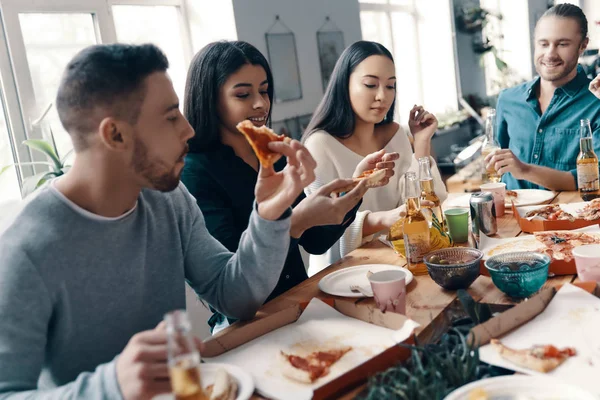 友達との夕食カジュアルウェアの若者のグループは 屋内でディナーパーティーを持っている間 ピザを食べて笑顔を身に着けている — ストック写真