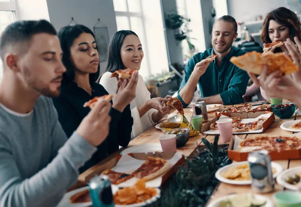 親友たちです カジュアルウェアの若者のグループは 屋内でディナーパーティーを持っている間 ピザを食べて笑顔を身に着けている — ストック写真