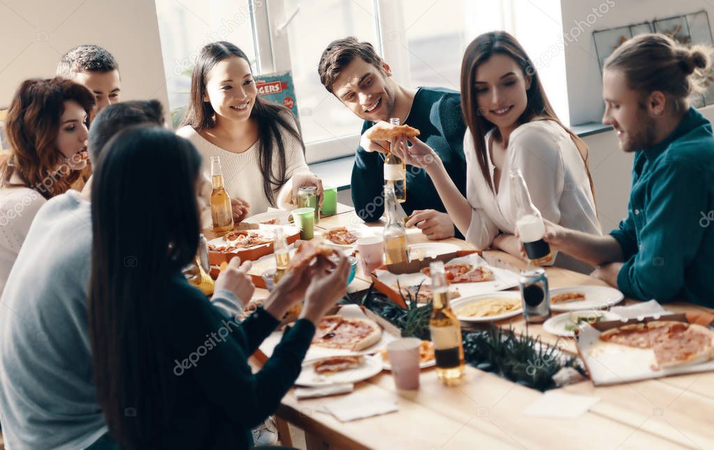 Group of young people in casual wear eating pizza and smiling while having a dinner party indoors               