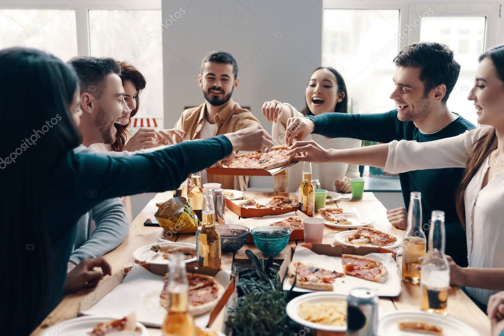 Who wants more pizza? Group of young people in casual wear picking pizza and smiling while having a dinner party indoors