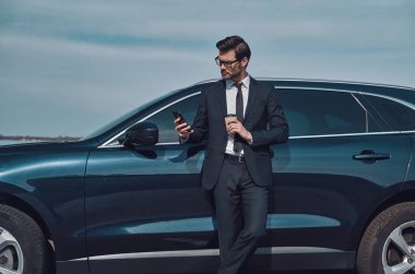 İş iletisi yazma. Yakışıklı genç işadamı akıllı telefon kullanarak ve açık havada arabasının yanında dururken kahve içme 