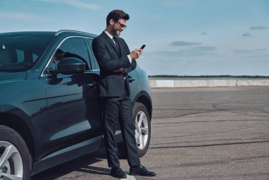 Kendine güvenen bir yönetici. Yakışıklı genç işadamı tam uzunlukta açık havada arabasının yanında dururken akıllı telefon kullanarak