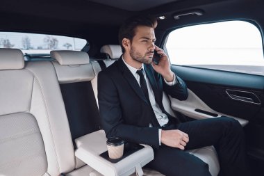 İş hareket halinde. Arabada otururken akıllı telefonda konuşurken tam takım elbiseli yakışıklı genç adam