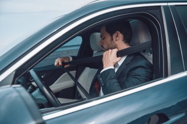 Güvenlik her şeyden önce gelir. Tam takım elbiseli yakışıklı genç adam araba sürerken emniyet kemeri uygulayarak