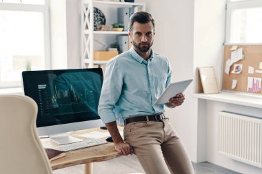 Genç ve başarılı genç ve modern iş adamı ofiste çalışırken kameraya bakıyor ve dijital tablet kullanıyor.