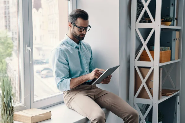 若い現代のビジネスマンは オフィスの窓の上に座っている間 デジタルタブレットを使用して作業 — ストック写真