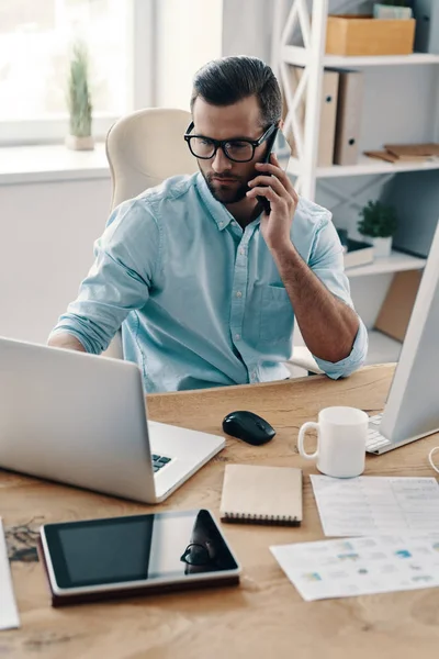 若い現代のビジネスマンが電話で話をしてオフィスに座っている間にコンピュータを使って — ストック写真