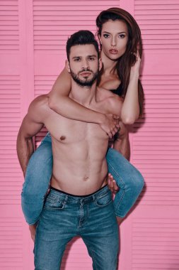 Yakışıklı gömleksiz adam pembe arka plan karşı dururken omuzlarında kız arkadaşı taşıyan
