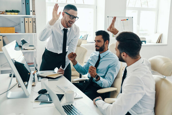 Group of young happy men in formalwear smiling and gesturing success in office 