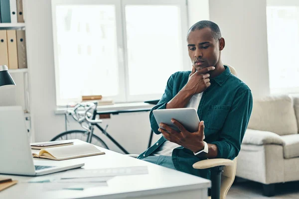物事を成し遂げるために急いでいる オフィスに座っている間 デジタルタブレットを使用して働くシャツの思慮深い若いアフリカの男性 — ストック写真