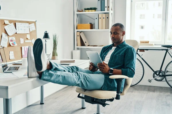 オフィスに座っている間 デジタルタブレットを使用してシャツを着て自信とスマートな思慮深い若いアフリカの男 — ストック写真