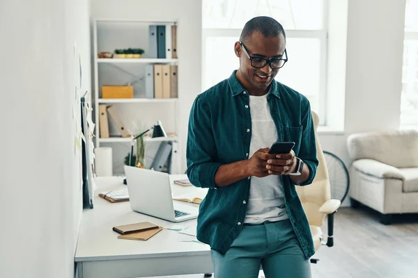 新しいモバイルアプリを使用して オフィスに立っている間 スマートフォンを使用してシャツを着たハンサムな若いアフリカ人男性 — ストック写真