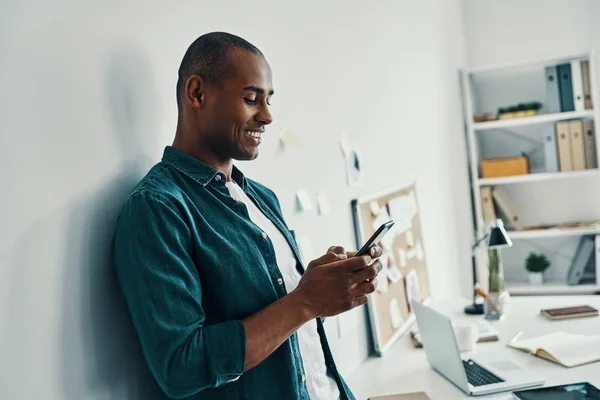 在手机上发短信 英俊的非洲青年男子穿着衬衫 站在办公室 使用智能手机 — 图库照片