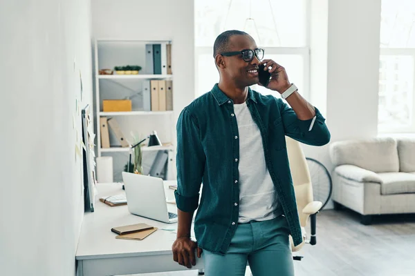 英俊的年轻非洲男子在衬衫上谈论智能手机和微笑 而站在家庭办公室 — 图库照片