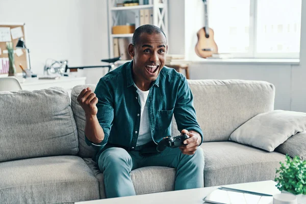 Winning game, Handsome young African man gesturing win and smiling while sitting on sofa in room