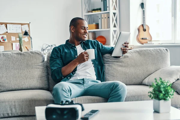 感觉舒适 英俊的年轻非洲男子使用数字平板电脑和微笑 而坐在室内拿着杯子和平板电脑 — 图库照片
