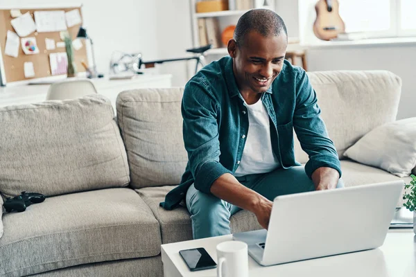 社交网络 英俊的年轻非洲男子使用笔记本电脑和微笑 而坐在房间里的陌生人的房间里的陌生人 — 图库照片
