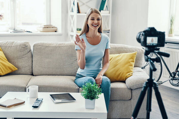 Beautiful young woman smiling and waving while making social media video at home    