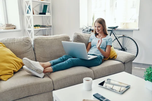 Güzel genç kadın gülümseme ile sallayarak ve evde kanepeüzerinde dinlenirken dizüstü bilgisayar kullanarak