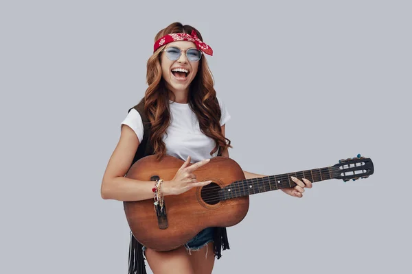 灰色の背景に立ちながらアコースティックギターを演奏し 笑顔で幸せなスタイリッシュな若い女性 — ストック写真