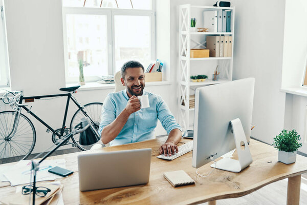 Handsome middle age man in smart casual wear using laptop and holding tea cup with smile 