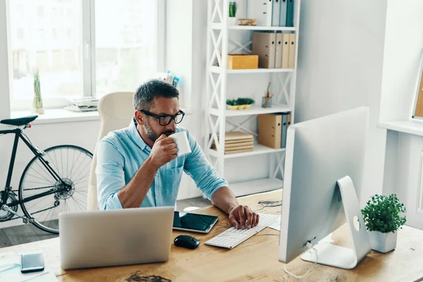 オフィスに座りながらコンピュータを使ってコーヒーを飲むスマートカジュアルウェアのハンサムな中年男性 — ストック写真