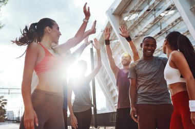 Spor giyimli bir grup genç birbirlerine beşlik kazandırıyor ve açık havada egzersiz yaparken gülümsüyor       