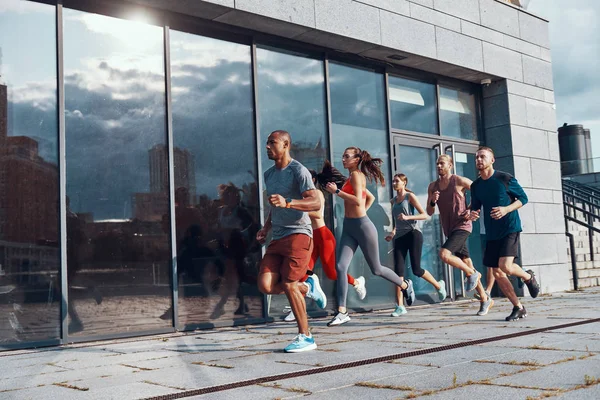 Group of people in sports clothing jogging outdoors in city at modern building with glass windows