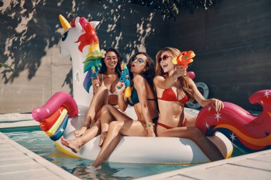 Mayo içinde çekici genç kadınlar gülümseyerek ve açık yüzme havuzunda şişme tek boynuzlu at üzerinde yüzerken fışkırtma tabancaları ile eğlenmek