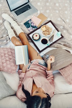 Pijamalı, elinde kitap ve gözlükleriyle evde dinlenen güzel genç bir kadın manzarası.          
