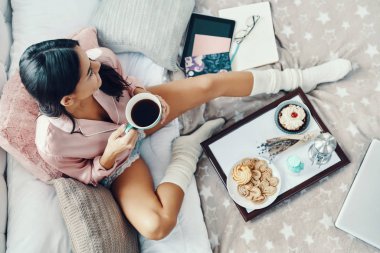 Pijamalı güzel bir genç kadın yatakta dinlenirken sıcak içkinin tadını çıkarıyor.   