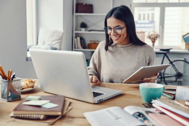 Dizüstü bilgisayar kullanan ve elinde dijital tablet tutan mutlu, güzel bir kadın.