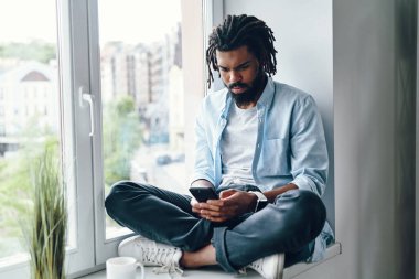 Pencerenin pervazında otururken akıllı telefon kullanan düşünceli bir Afrikalı.                    