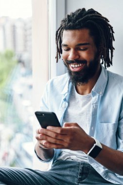 Akıllı telefon kullanan ve pencerenin pervazında otururken gülümseyen yakışıklı bir Afrikalı.            