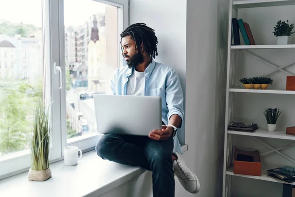 窓の上にまだ屋内に座っている間 ノートパソコンを使用して離れて見てハンサムな若いアフリカ人男性 — ストック写真
